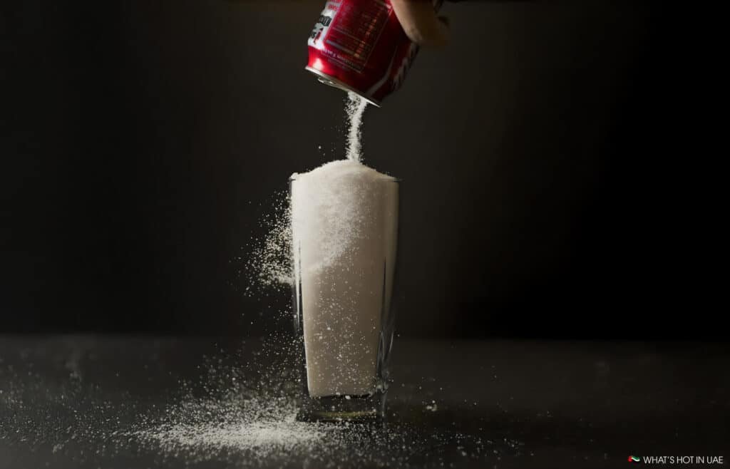A hand pours sugar from a red soda can into a tall glass, overfilling it with sugar and spilling onto a dark background.