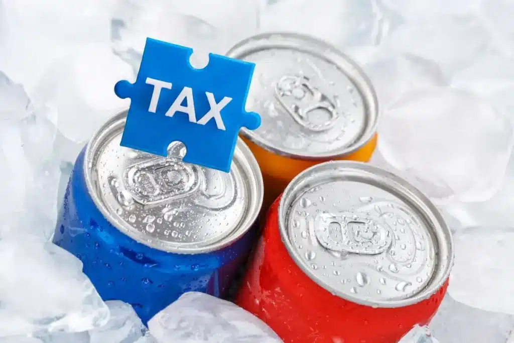 Three soda cans in different colors are surrounded by ice, with a blue puzzle piece labeled 'TAX' placed on top of them. UAE Sugar Tax