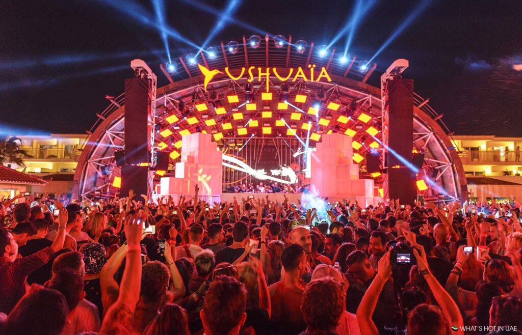 A large crowd enjoys a vibrant concert at Ushuaïa, with a brightly lit stage and colorful lighting effects overhead.