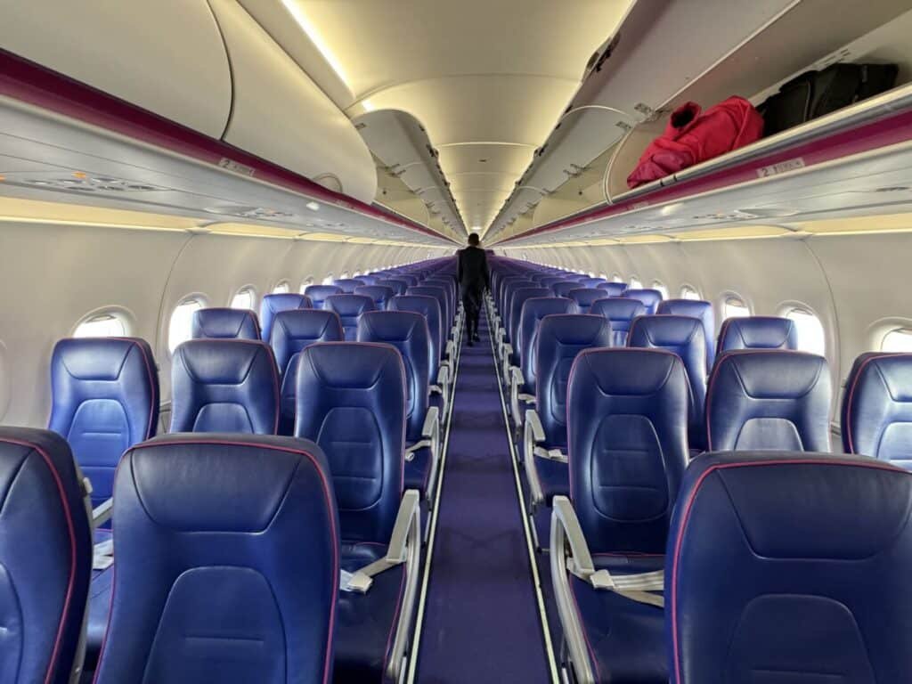 Interior of an empty Wizz Air Abu Dhabi flights airplane cabin with rows of blue seats, overhead compartments open with some luggage inside, and a single person walking down the aisle.