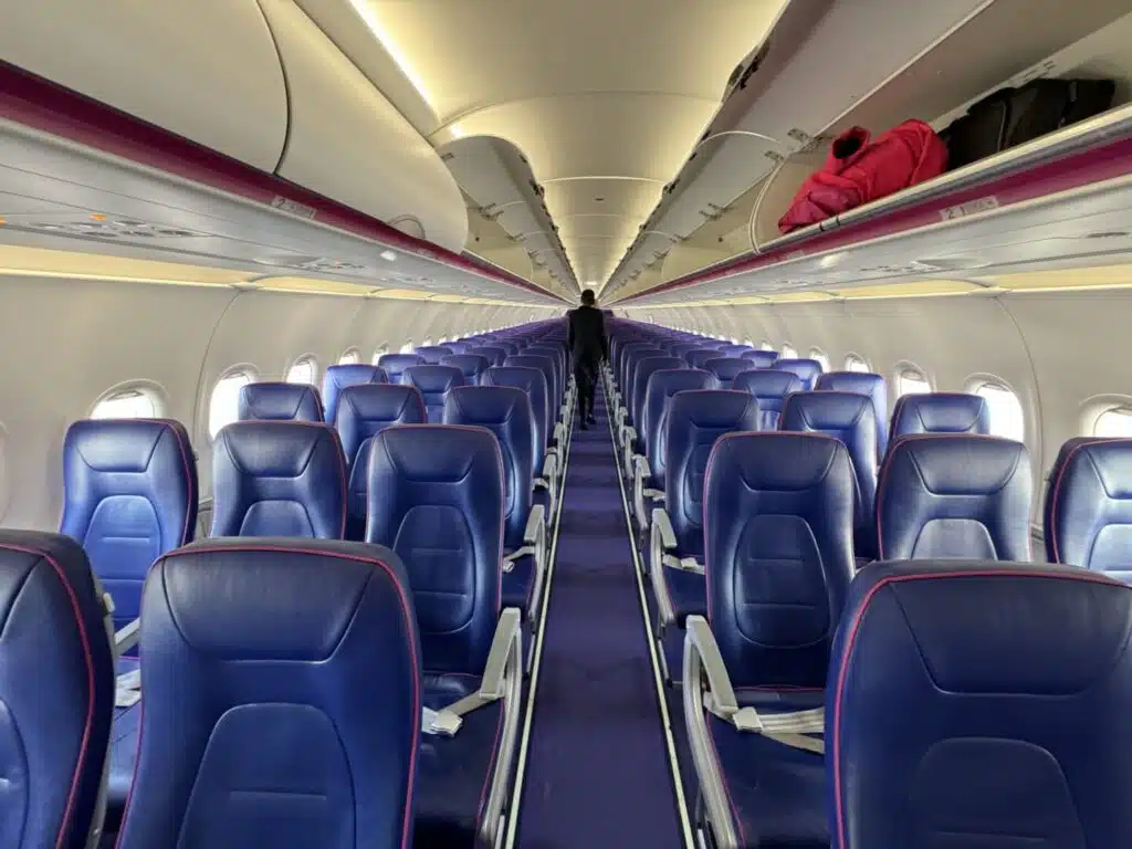 Interior of an empty Wizz Air Abu Dhabi flights airplane cabin with rows of blue seats, overhead compartments open with some luggage inside, and a single person walking down the aisle.