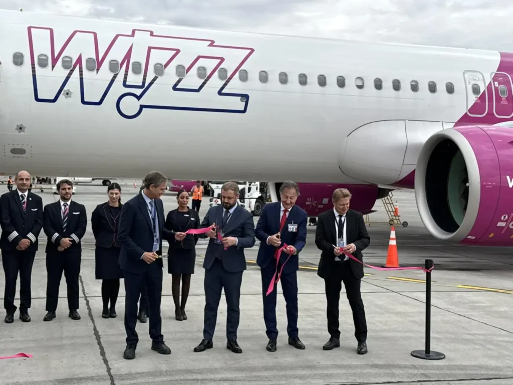 A group of people in formal attire stand in front of a Wizz Air Abu Dhabi flights airplane during a ribbon-cutting ceremony on an airport tarmac.
