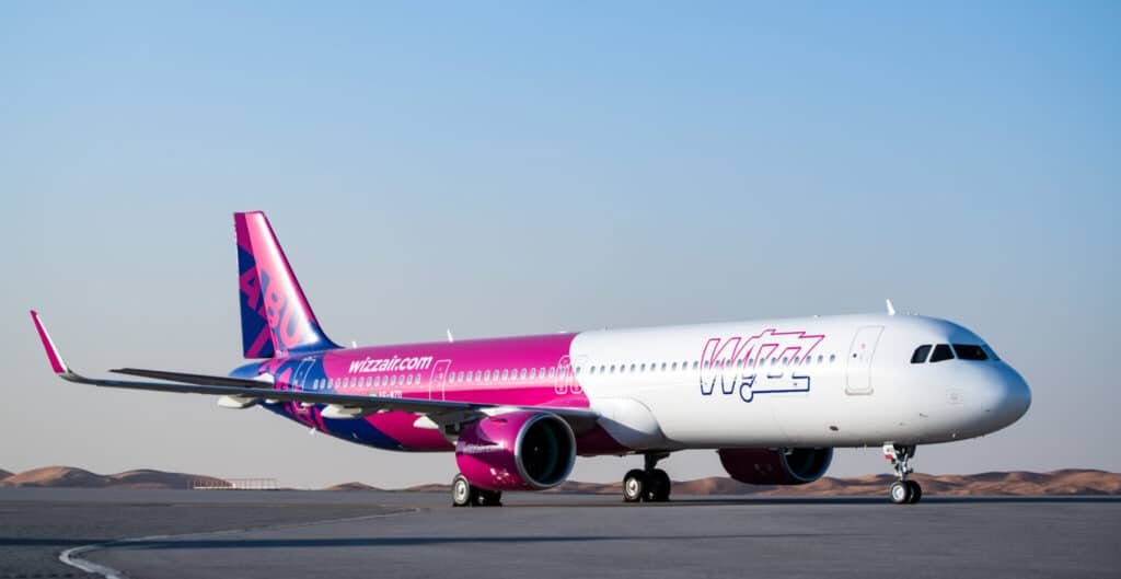 A commercial aircraft with a pink and purple livery, featuring the Wizz Air branding, is taxiing on an airport runway against a clear blue sky.