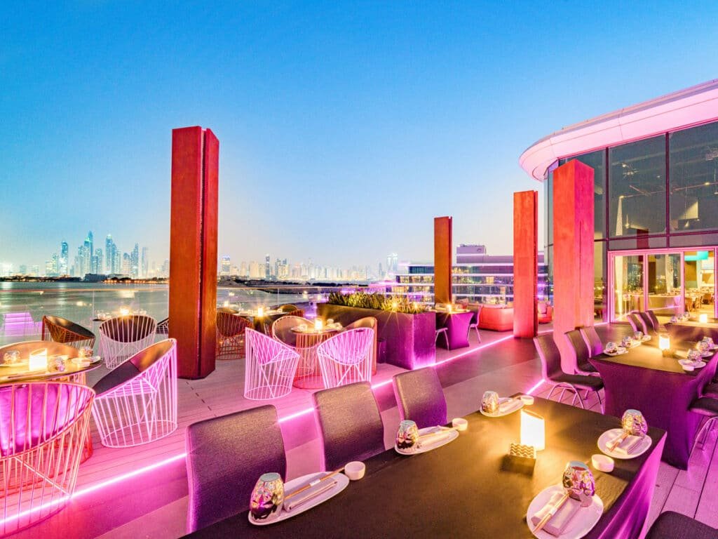 A rooftop restaurant with pink lighting, featuring modern dining tables and chairs, offering a view of a city skyline across a body of water during sunset. Japanese Restaurants Date Night.