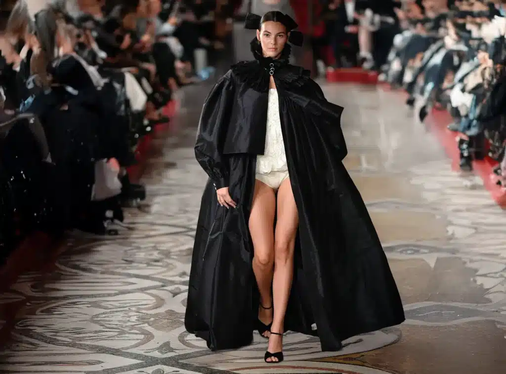 A model walking down a runway wearing a dramatic black cape with puffed shoulders, over a white outfit, and black heels. The setting is an ornate, crowded fashion show venue.