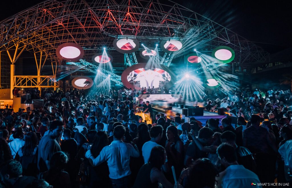 A crowded indoor nightclub scene with bright stage lights and circular structures hanging from the ceiling, illuminating a lively crowd of people dancing and socializing under a large, open metal frame structure.
