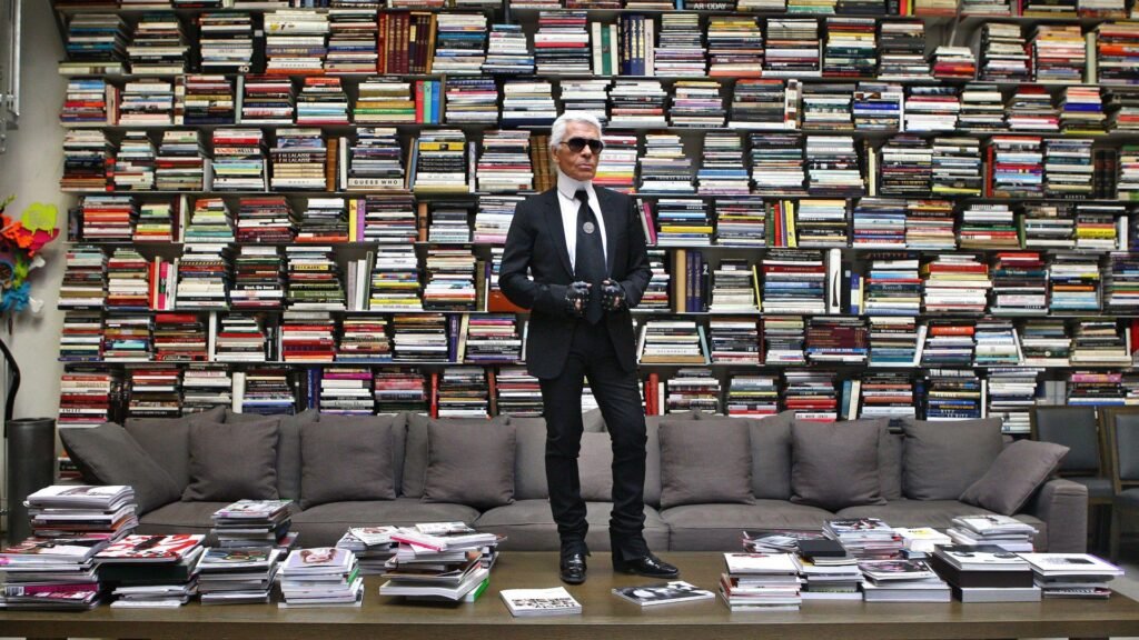 A person dressed in a black suit, sunglasses, and gloves, stands on a coffee table in front of a large wall filled with books. There is a gray couch behind the table, and stacks of magazines are neatly arranged on the table's surface.