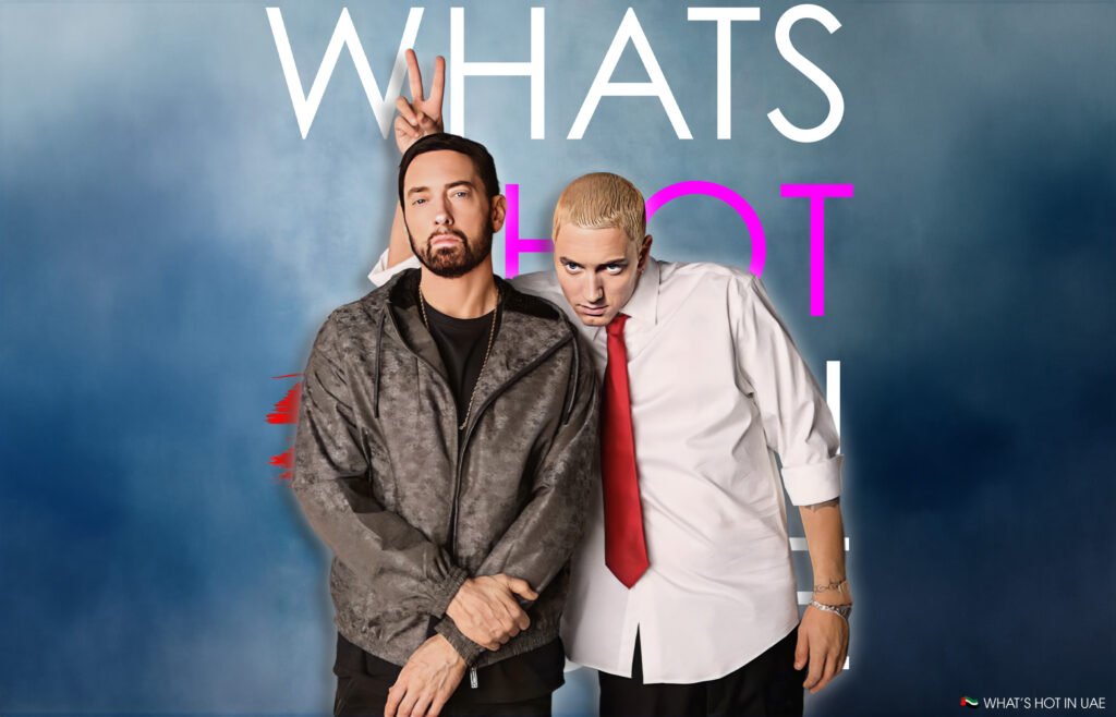 The 2025 Rap Drought: Eminem posing in front of a backdrop with text reading 'WHAT'S HOT.' One person, wearing a gray jacket, stands with arms crossed while the other, wearing a white shirt and red tie, leans in with a playful gesture.
