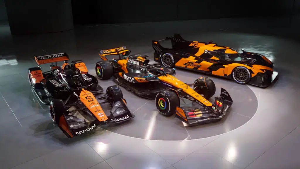Three racing cars with similar orange and black liveries are displayed on a sleek Sotheby’s showroom floor. The cars are designed for different racing series, with varying aerodynamic features and sponsorship logos. The scene is well-lit, highlighting the shiny surfaces and detailed designs of the vehicles.