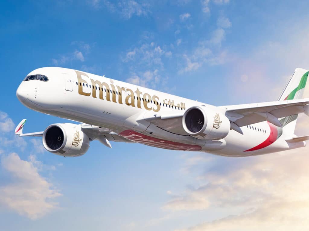 An Emirates airplane flying against a blue sky with scattered clouds, featuring the airline's logo and colors on its fuselage and tailfin.