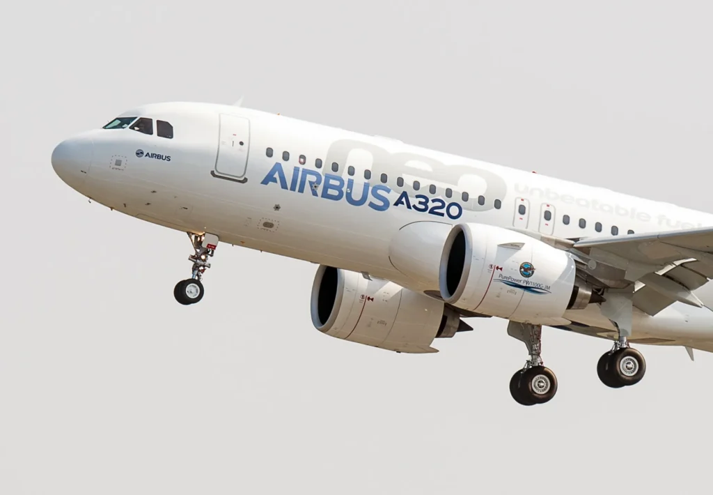A close-up view of an Airbus A320 aircraft in flight, showing its landing gear extended and displaying the Airbus logo and model name on the fuselage.