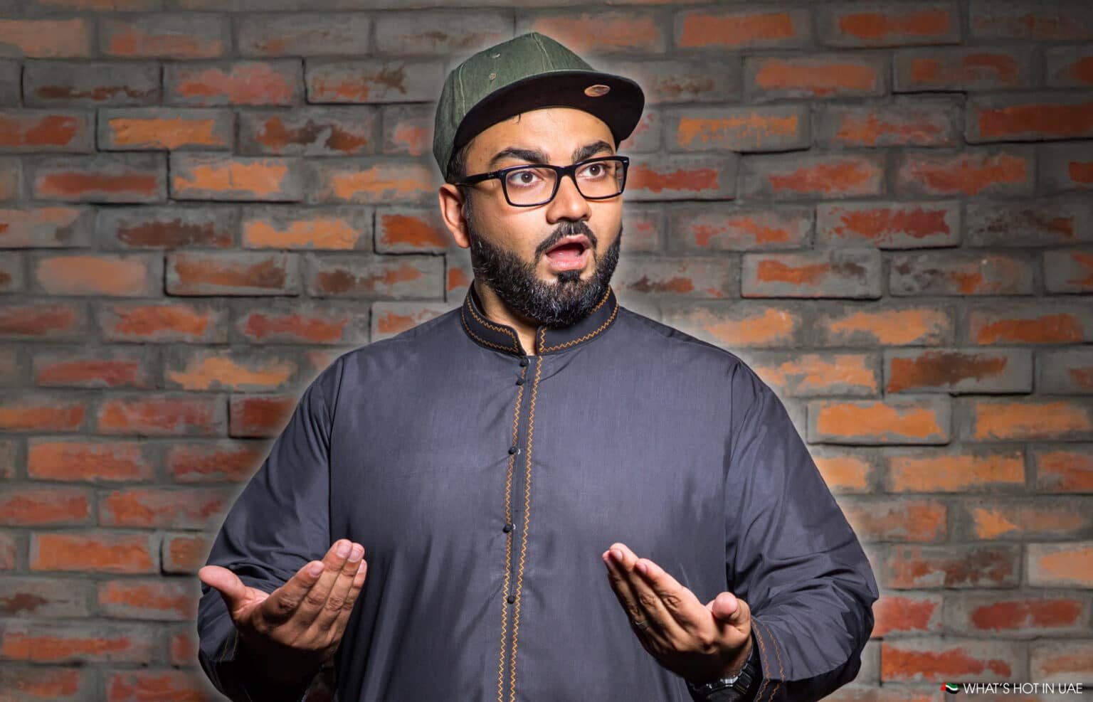 A man wearing a dark traditional outfit and a cap, with an expressive facial expression, stands in front of a brick wall background.