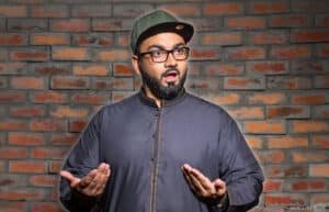 A man wearing a dark traditional outfit and a cap, with an expressive facial expression, stands in front of a brick wall background.