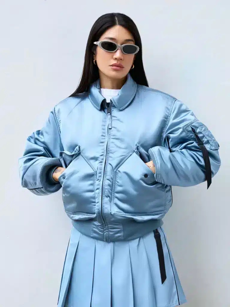 Peggy Gou wearing a shiny light blue bomber jacket and matching pleated skirt, accessorized with small oval sunglasses and gold hoop earrings, posing against a light-colored wall.