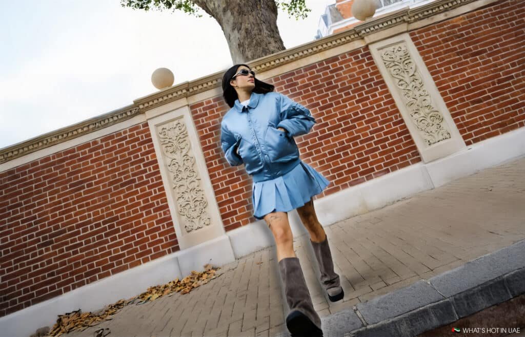 A woman wearing a light blue jacket and skirt with knee-high gray boots walks past a brick wall with decorative stone carvings, under a tree in an urban setting.