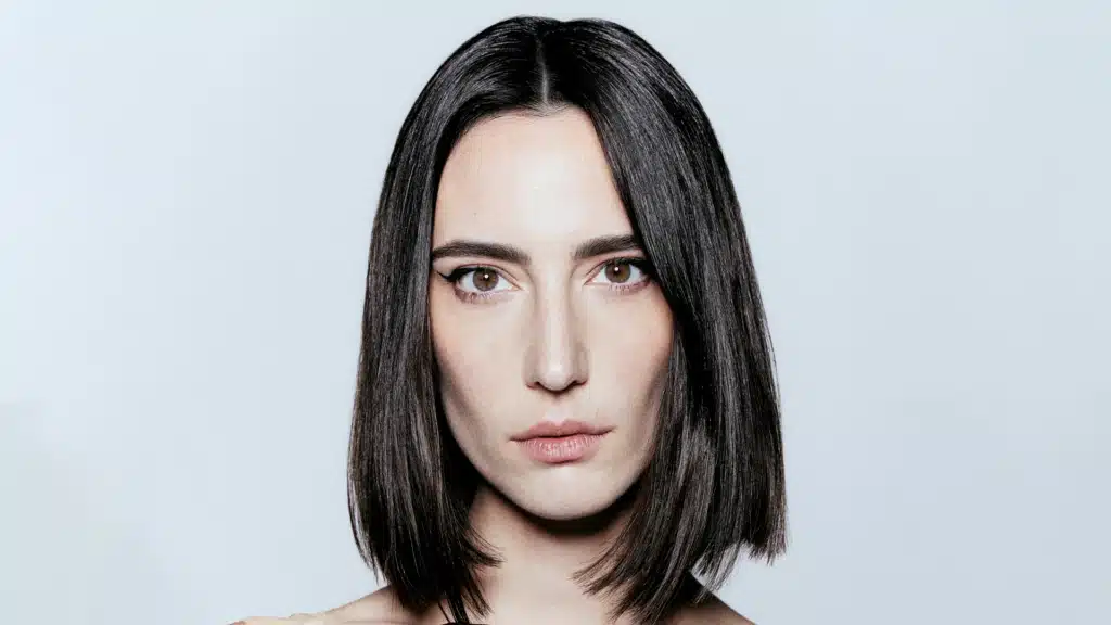 Amelie Lens with straight dark hair and intense eyes, looking directly at the camera against a plain background.