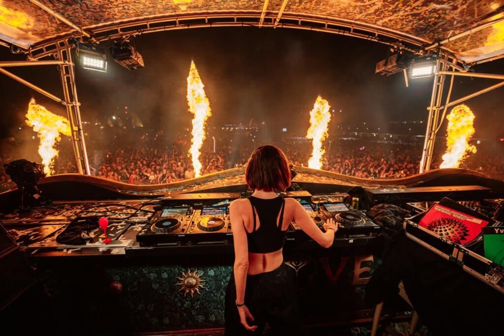 A DJ is performing on stage at a music festival, with flames shooting into the air in front of a large, enthusiastic crowd at night.