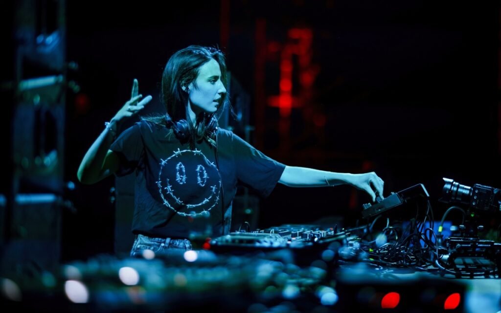 Amelie Lens wearing a black t-shirt with a smiley face graphic is performing on stage, adjusting equipment under vibrant lighting.