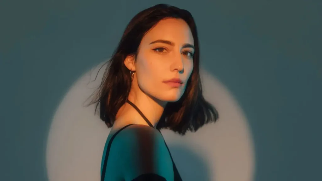 A woman with shoulder-length dark hair, wearing a black top, is looking over her shoulder with a serious expression. The image is artistically lit with a spotlight effect against a teal background, highlighting her face and silhouette.