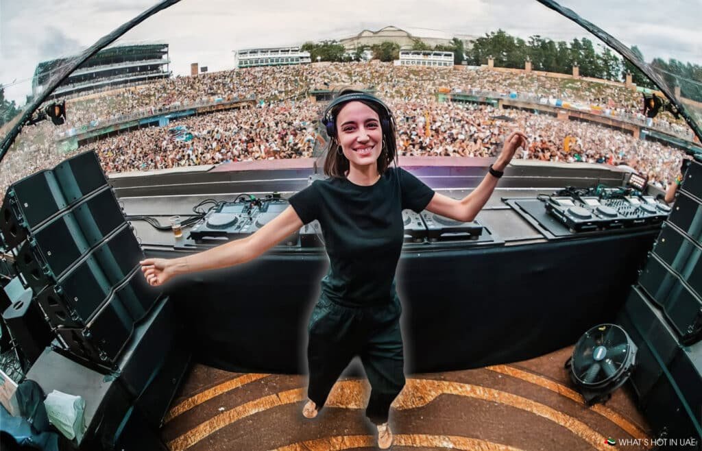 A DJ wearing black clothing and headphones smiles and dances at a large outdoor music festival, with a massive crowd cheering in the background.