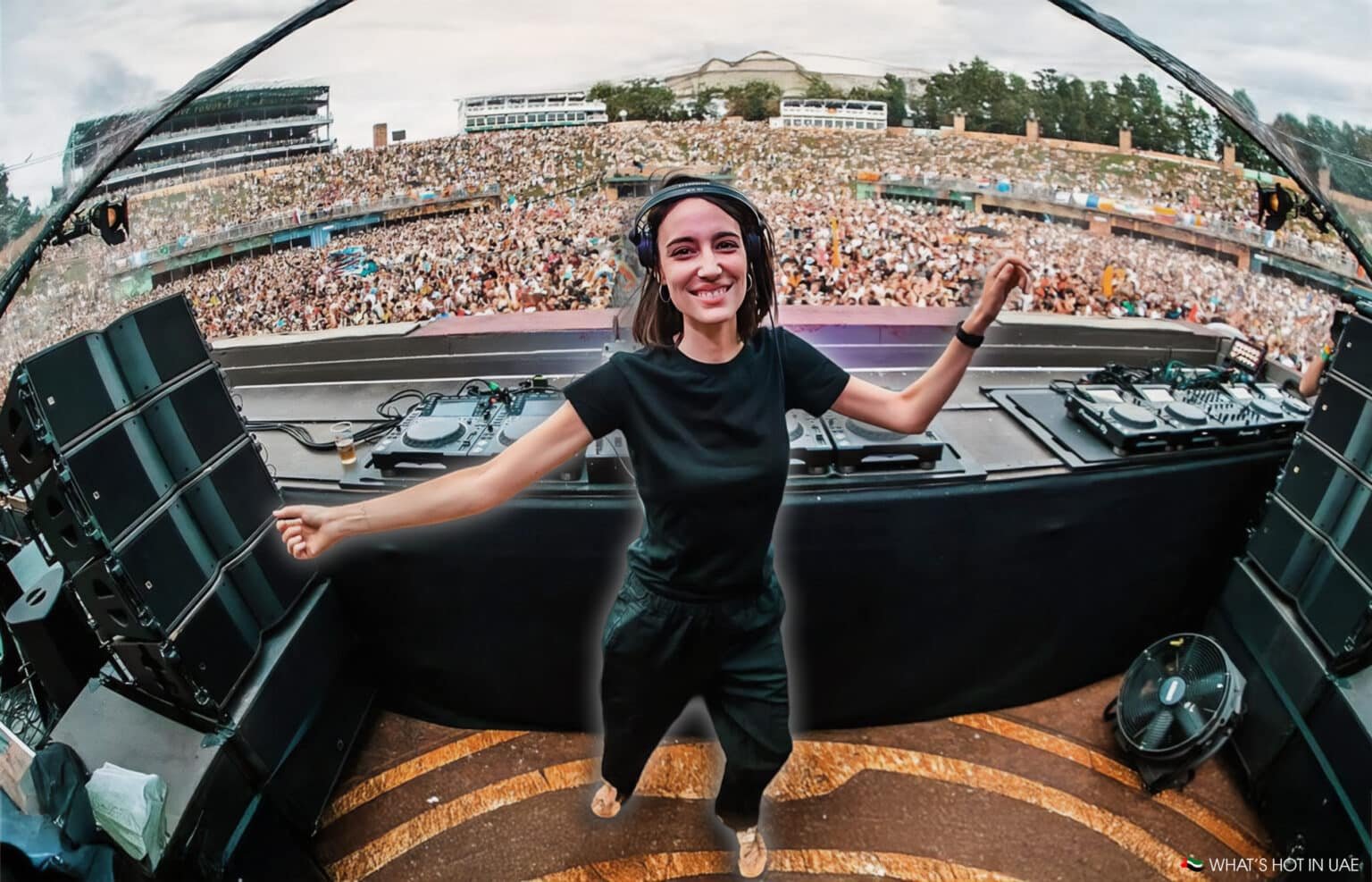A DJ wearing black clothing and headphones smiles and dances at a large outdoor music festival, with a massive crowd cheering in the background.