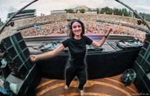A DJ wearing black clothing and headphones smiles and dances at a large outdoor music festival, with a massive crowd cheering in the background.