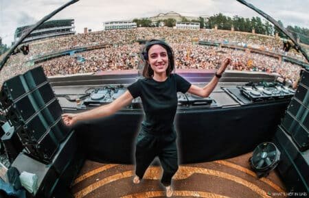 A DJ wearing black clothing and headphones smiles and dances at a large outdoor music festival, with a massive crowd cheering in the background.
