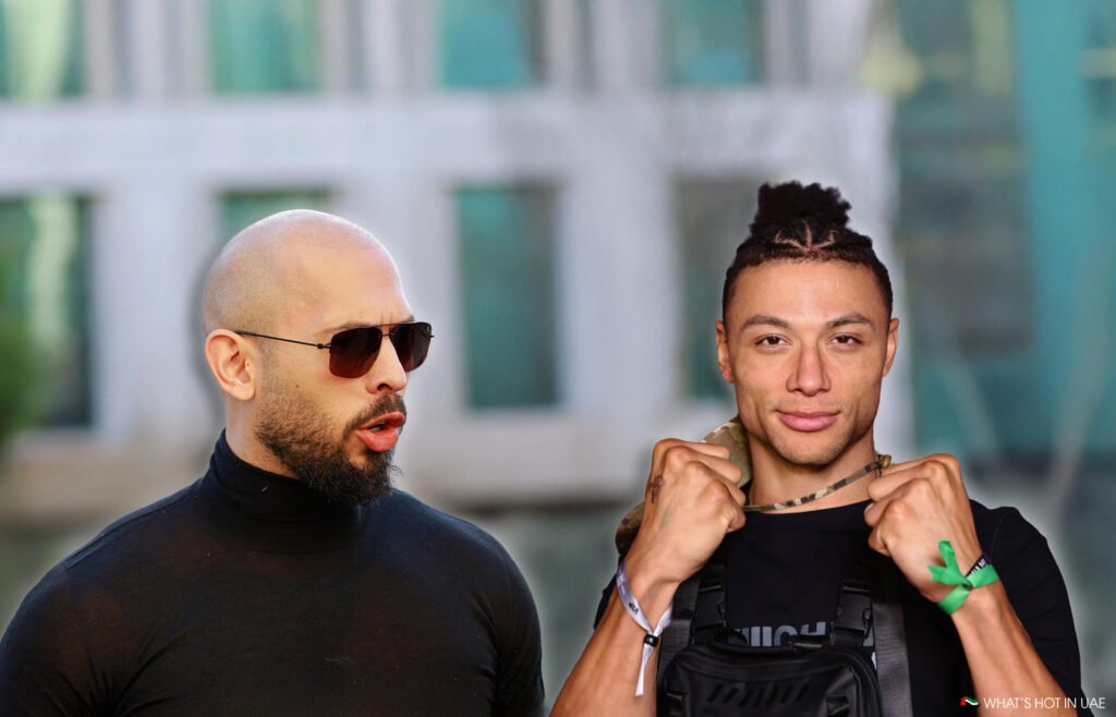 Two men standing outdoors; one wears a black turtleneck and sunglasses, while the other has a braided hairstyle and holds a camo pattern strap.