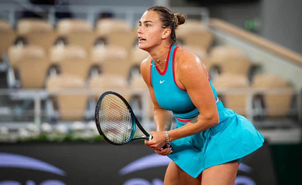A tennis player in a blue outfit prepares to hit a ball on an indoor court, with empty seats in the background.