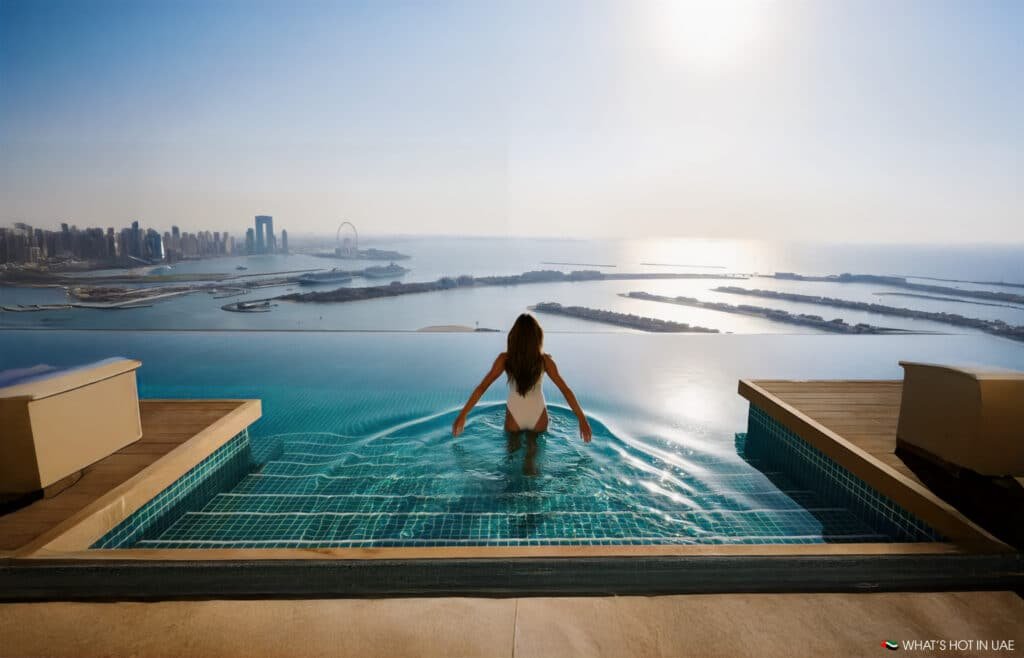A woman in a white swimsuit sits at the edge of an infinity pool, overlooking a sprawling cityscape and waterfront in the distance, under a clear blue sky.