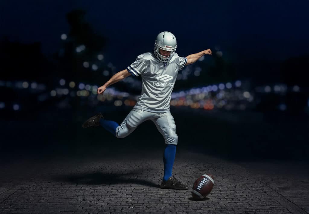 A football player in full gear, including a helmet and blue socks, is in the motion of kicking an American football on a cobblestone surface at night, with blurred city lights in the background.