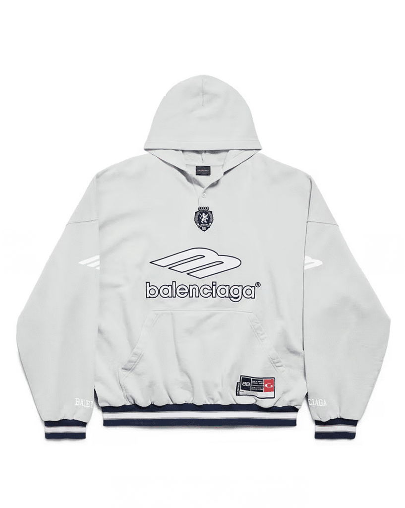 A gray Balenciaga hoodie featuring a front pocket and various logos, including a crest and a stylized "m", with contrasting navy stripes on the hem and cuffs.