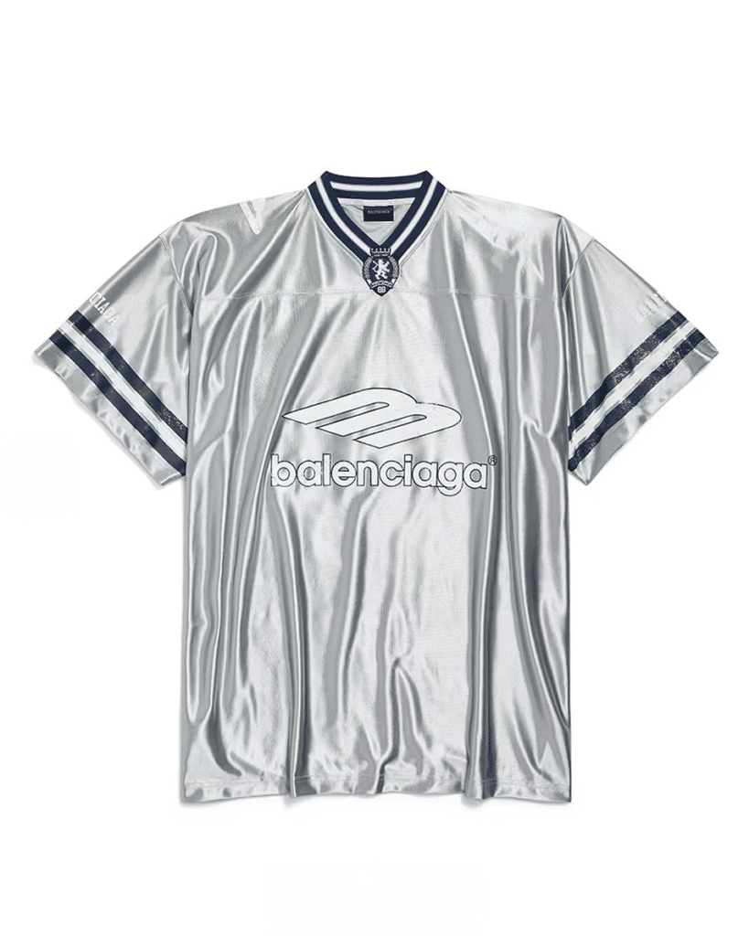 A silver jersey with a shiny finish, featuring dark blue stripes on the sleeves, a dark blue collar, and the word 'balenciaga' printed on the front with a logo above it.