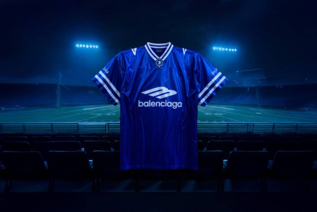 A blue sports jersey with white stripes and 'Balenciaga' branding, displayed in front of an empty stadium under bright lights. Balenciaga Football Series