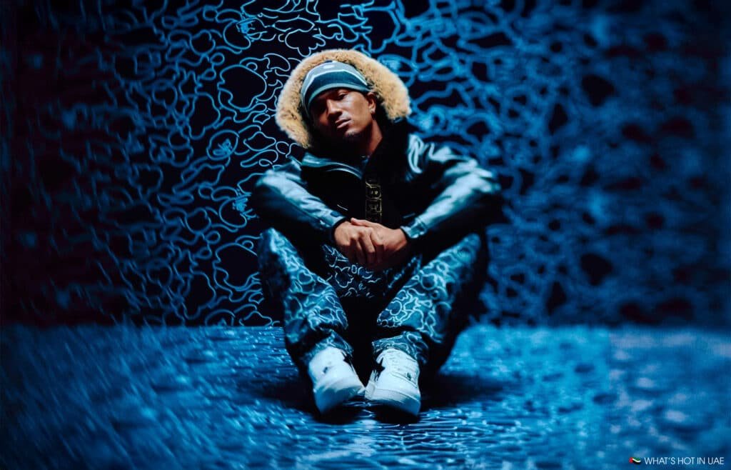 A person wearing a fur-lined hooded jacket and camo pants sits with crossed arms on a patterned blue floor and wall backdrop. The environment has a swirling blue pattern, creating a moody, abstract atmosphere.