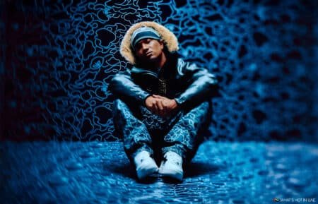 A person wearing a fur-lined hooded jacket and camo pants sits with crossed arms on a patterned blue floor and wall backdrop. The environment has a swirling blue pattern, creating a moody, abstract atmosphere.
