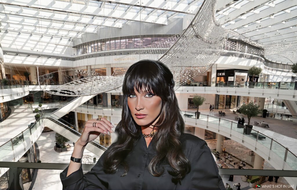 Bella Hadid in Dubai with long dark hair and wearing a black outfit stands in a brightly lit, spacious shopping mall with multiple levels and modern architectural design, featuring a large decorative installation hanging from the ceiling.