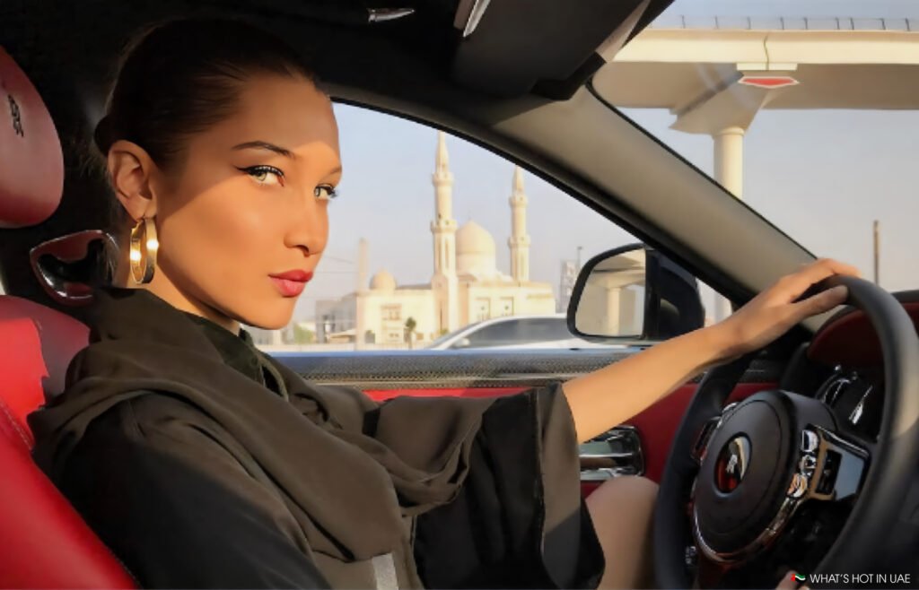 Bella Hadid in Dubai with large earrings sitting in the driver's seat of a luxury car with a mosque visible through the window.