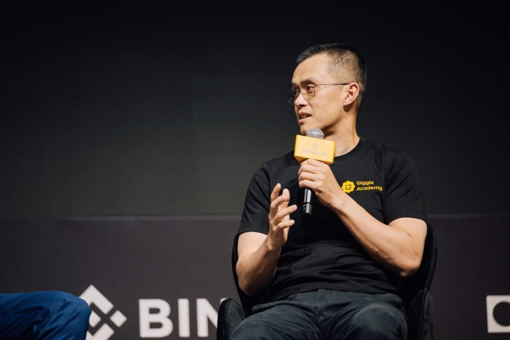 CZ sitting on a stage at Binance Blockchain Week 2025, holding a microphone with a Binance logo, wearing a "Giggle Academy" t-shirt.