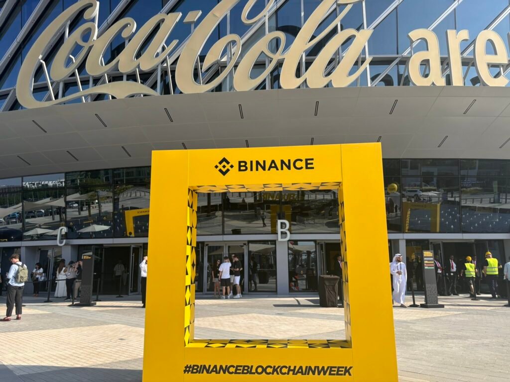 Entrance of Coca-Cola Arena featuring a large yellow Binance frame promoting Binance Blockchain Week 2025, with people standing in the foreground.