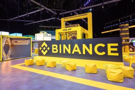 A large exhibition booth featuring the Binance logo prominently displayed in yellow and black. The booth includes decorative elements, seating with yellow bean bags, and signs promoting Binance Blockchain Week.