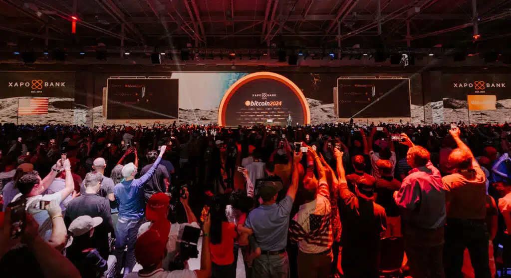 A large crowd of people facing a stage with a presentation screen displaying the words 'Bitcoin 2024,' with the Xapo Bank logo visible. The audience is using phones and cameras to capture the event inside a spacious conference hall with a high ceiling.