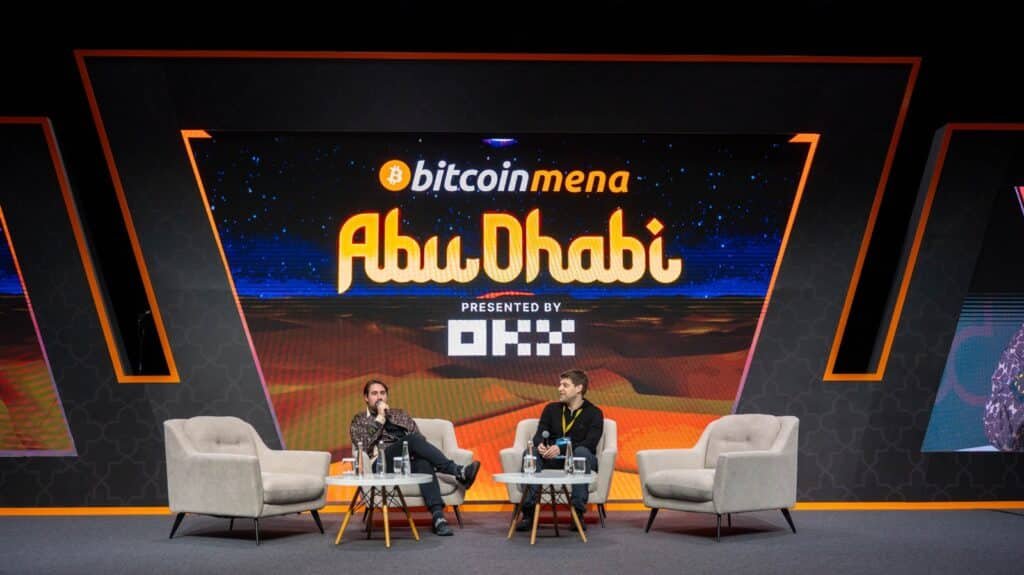 Two speakers seated on stage at the Bitcoin MENA Abu Dhabi event, with signage in the background. The stage features modern furniture and event branding, emphasizing technology and finance themes.