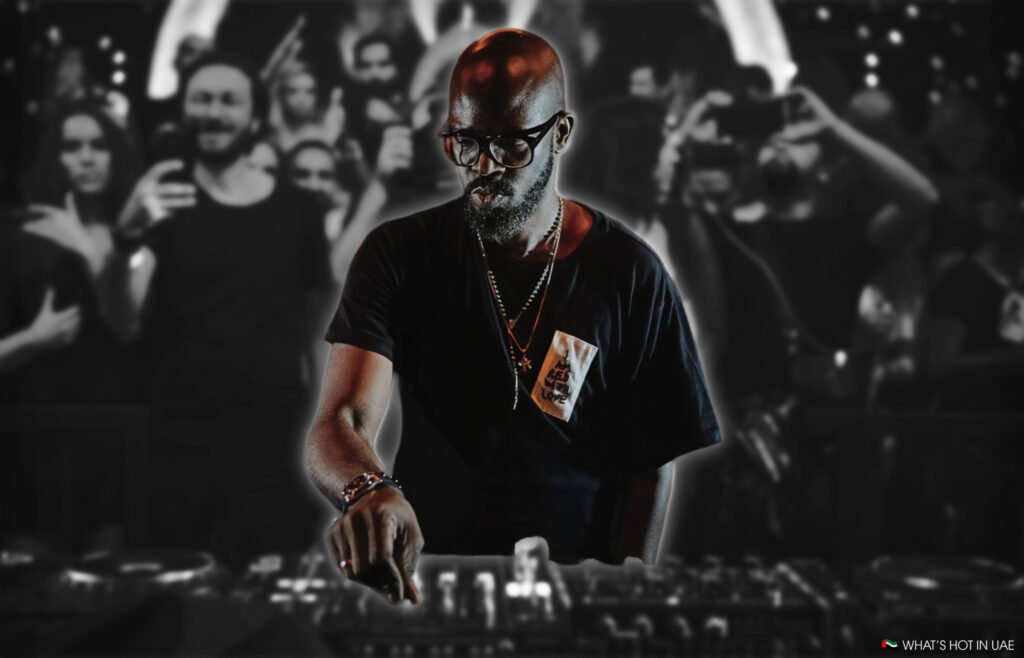 Black Coffee At Ushuaïa Experience wearing a black t-shirt and glasses, focused on mixing music at a DJ console with a blurred audience in the background.