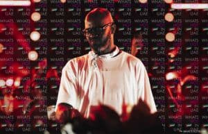 A man in a white shirt and bandana stands in front of a backdrop with 'What's Hot in UAE' text and UAE flags, surrounded by circular lights.