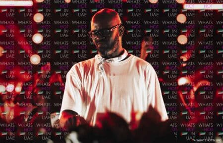 A man in a white shirt and bandana stands in front of a backdrop with 'What's Hot in UAE' text and UAE flags, surrounded by circular lights.