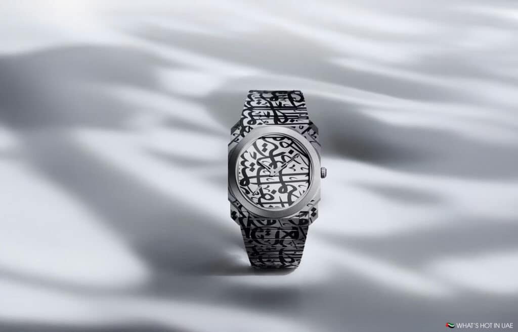 A luxury wristwatch with a silver case and band, featuring intricate black Arabic calligraphy design on both the dial and the strap, set against a blurred gray background.