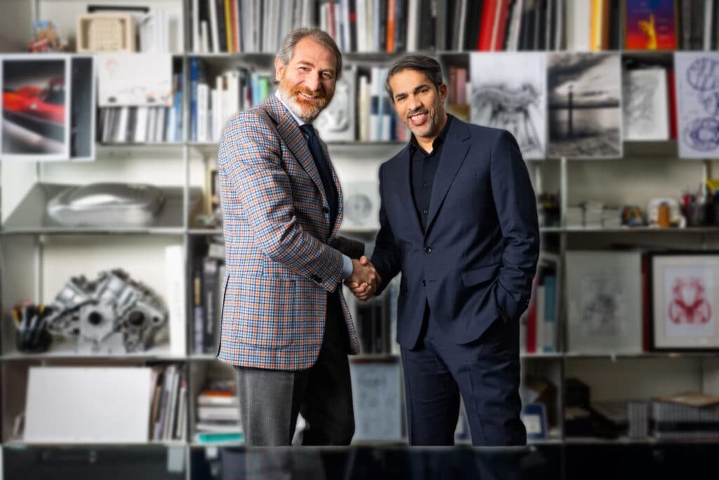 Two men in suits shaking hands in front of a bookshelf filled with books and sketches, indicating a professional setting.