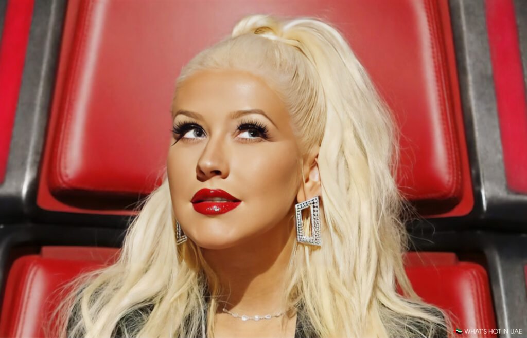 A woman with long blonde hair styled in a high ponytail, wearing red lipstick, large square earrings, and a black outfit, sits in a red chair and looks upward.