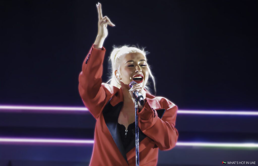 Christina Aguilera in Abu Dhabi with blonde hair, wearing a red blazer, sings into a microphone under bright stage lights.